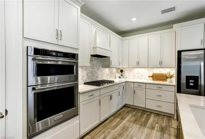 Kitchen with light hardwood / wood-style floors, tasteful backsplash, custom exhaust hood, white cabinets, and appliances with stainless steel finishes